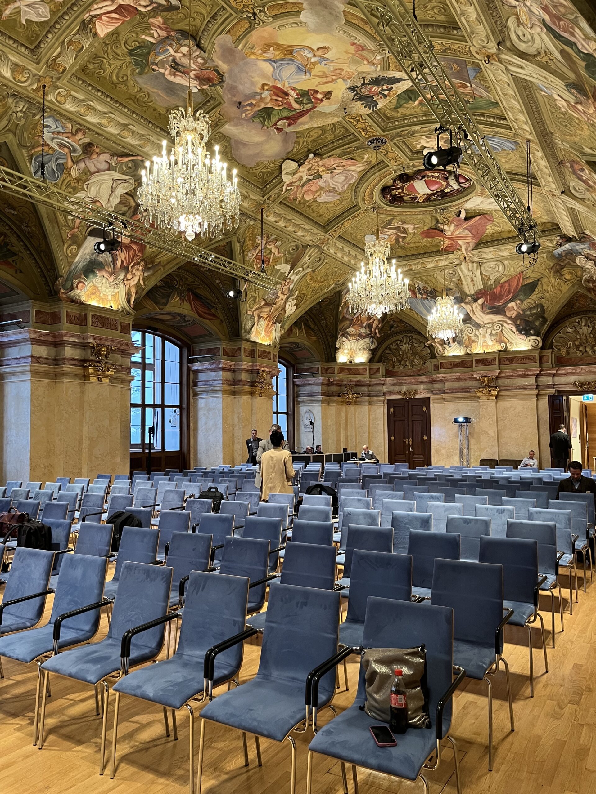 View of the venue room at the baroque Palais Niederösterreich in the Circular Carbon Economy Summit in Vienna 2024. The painted, golden, baroque ceiling has two big chandeliers. On the floor there are several rows of comfortable blue chairs for the audience.