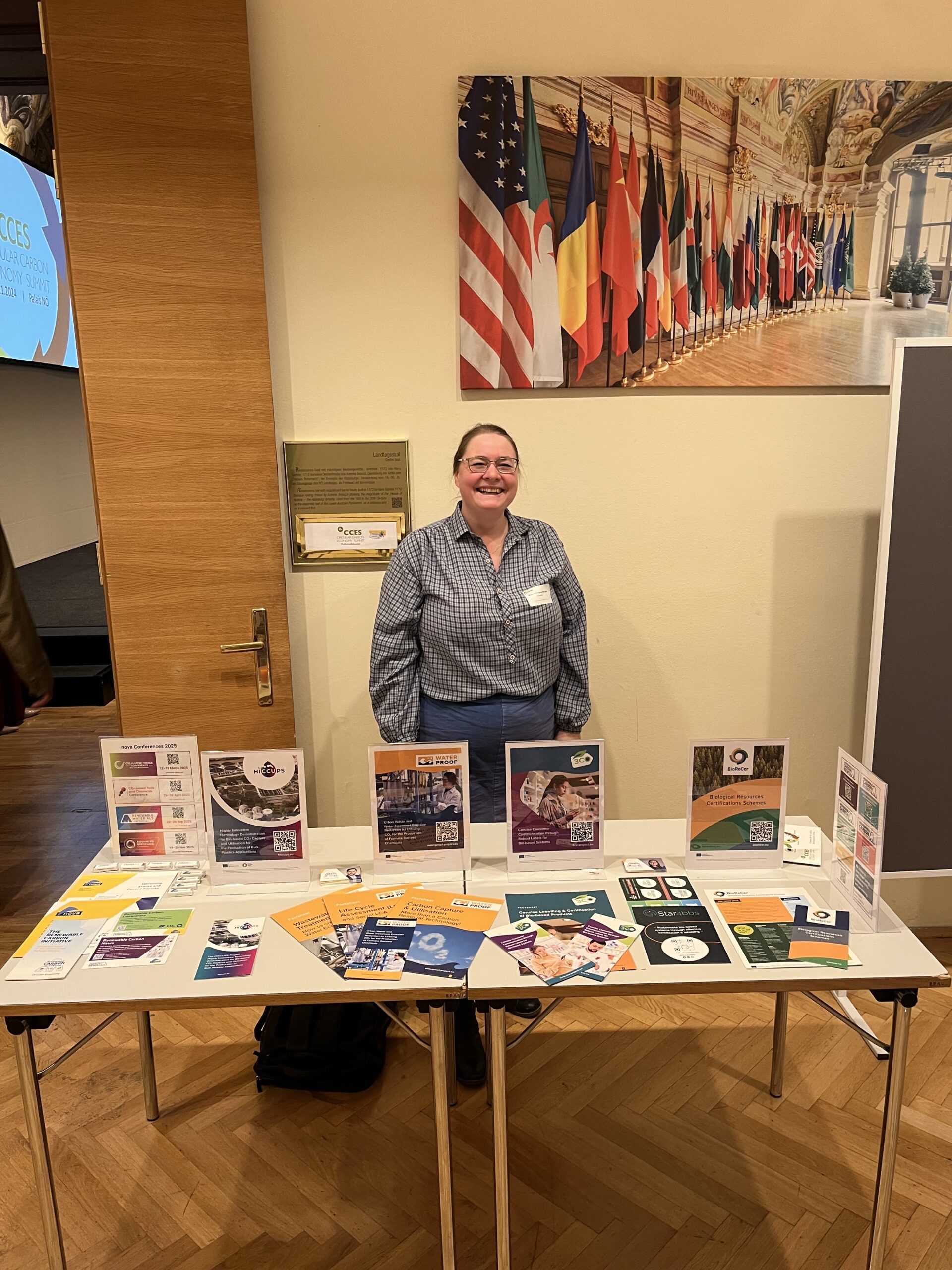 Anke Schwarzenberger in front of the table showing information material of several Horizon Europe projects at the Circular Carbon Economy Summit in Vienna. Anke wears a blue blouse and a blue skirt.