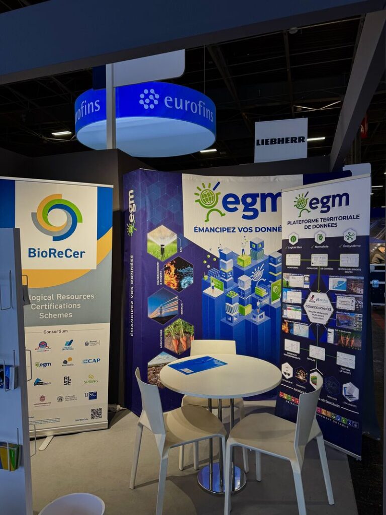 A conference booth in a dark hall. A round, white table with three chairs stands in front of three roll-up banners. The two left ones presenting the BioReCer project partner gem, the left one is the official BioReCer roll-up banner. All roll-ups are kept mostly in blue, green, yellow and gray.