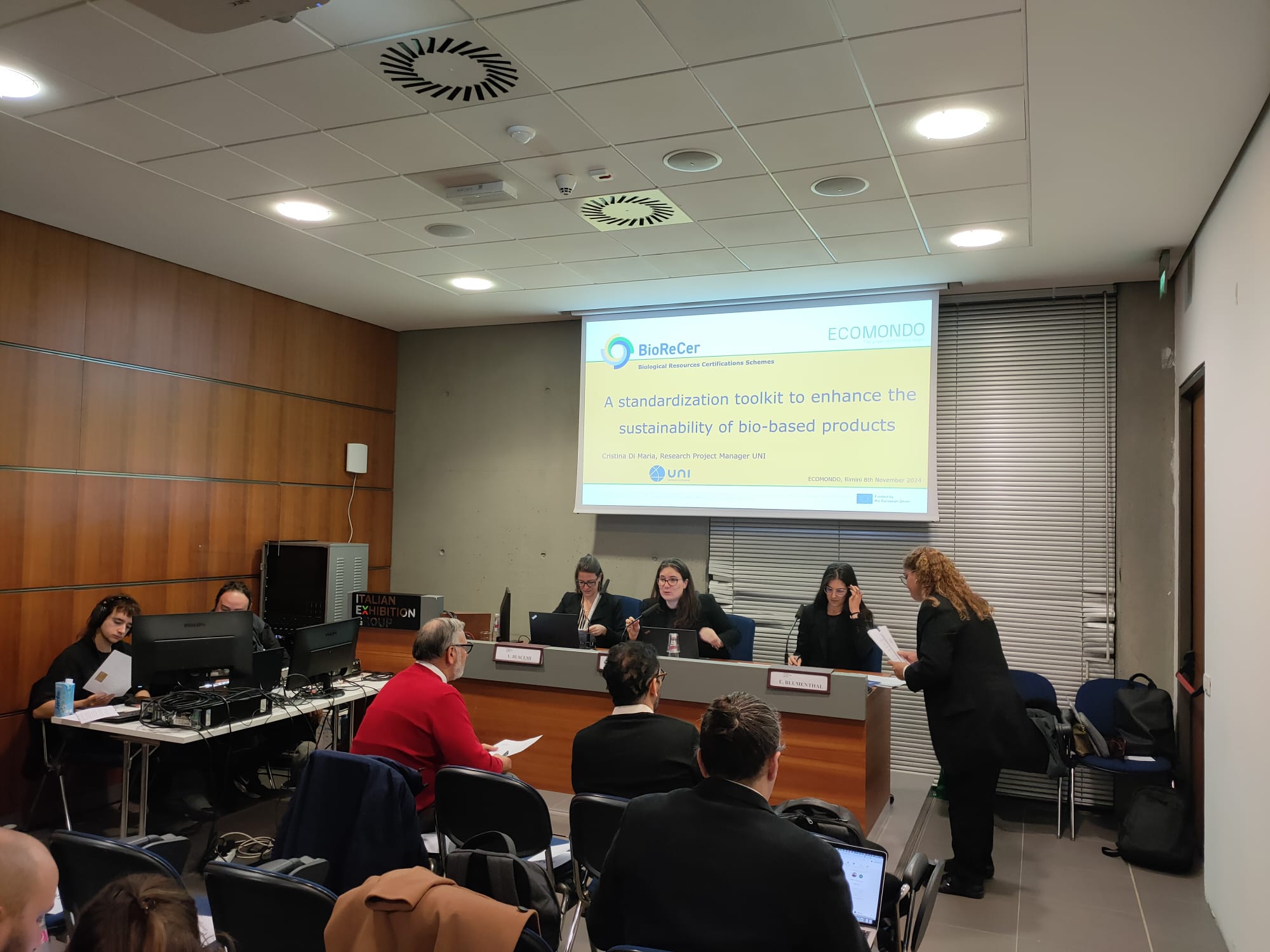 Ecomondo workshop by UNI. View into the lecture room with a small audience and the three workshop organisers sitting in front of a screen facing the participants. It is a small room with a wooden left facade.