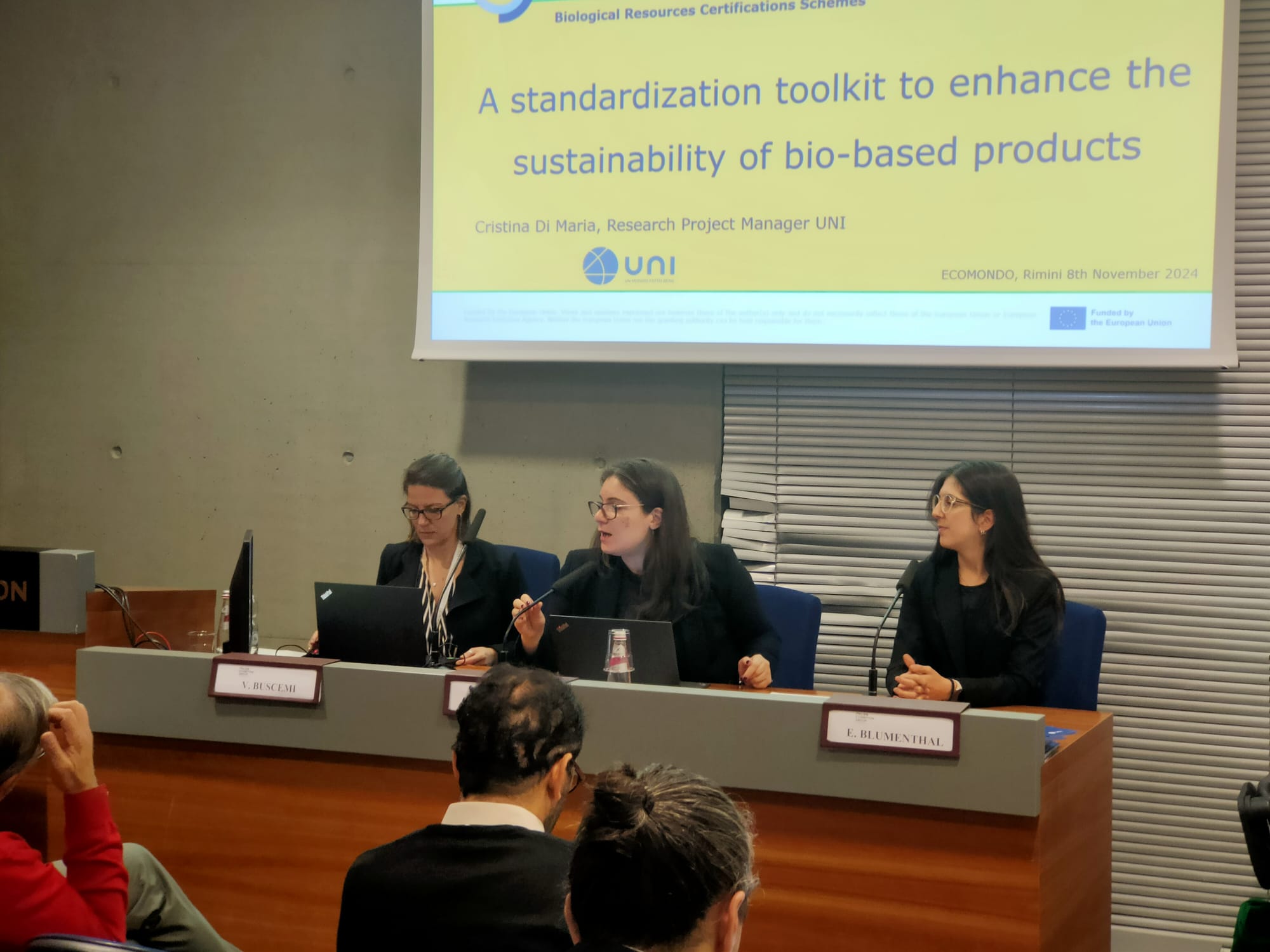 The three workshop organisers facing the workshop participants at Ecomondo in Rimini, Italy. The three Italian ladies from UNI, Italy, sit in front of a screen facing the audience. On the left is V. Buscemi, in the middle Federica Fugaroli and on the right is Elisa Bumenthal.